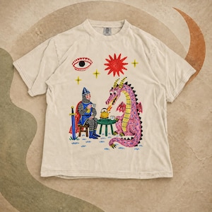 May include: A beige short-sleeve t-shirt featuring a graphic print of a knight in chainmail armor sitting on a stool and a pink dragon drinking tea together. Above them are a red sun, a single eye, and yellow stars. A blue sword stands nearby.