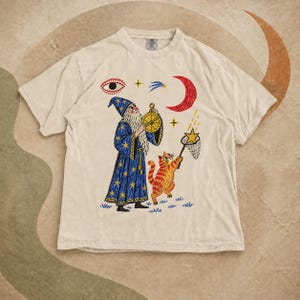 May include: A beige short-sleeve t-shirt featuring a graphic print of a wizard in a blue star-patterned robe holding an astrolabe, accompanied by an orange tabby cat catching a yellow star with a net under a red crescent moon and an eye.