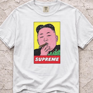 May include: White t-shirt featuring a pop art style illustration of a person smoking a cigar, set against a yellow background. The word "SUPREME" is displayed in red block letters at the bottom. The shirt has a Comfort Brand tag.