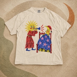 以下が含まれることがあります： ベージュの半袖Tシャツ。赤いローブを着た黄色い太陽が青い角笛を吹き、赤いナイトキャップを被った黄色い三日月が赤いベッドで寝ている様子が描かれたグラフィックプリント。ベッドにはピンクと黄色の星が散りばめられた青い毛布が掛けられている。