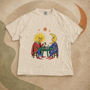 Könnte beinhalten: Ein beiges Kurzarm-T-Shirt mit einem Grafikdruck von Sonne und Mond, die an einem Tisch Schach spielen. Die Sonnenfigur trägt ein rotes Gewand, die Mondfigur ein blaues Gewand mit Sternen. Das Design zeigt zudem kleine Sterne und einen großen Stern.