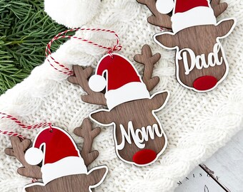 May include: Three wooden Christmas reindeer ornaments, each with a Santa hat. The ornaments are inscribed with the names "Dad", "Mom", and "Ashley". They are brown, red, and white, with red and white string for hanging.