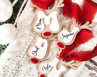 May include: Four wooden reindeer ornaments with red noses and white painted areas. The ornaments are personalised with the names "Mom", "Dad", "Jasmine", and "Nyla".