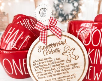 May include: A wooden gingerbread cookie recipe ornament with a red and white checkered ribbon. The ornament is round and has a gingerbread man illustration. The recipe is written in a handwritten font and includes ingredients like soft butter, brown sugar, molasses, flour, baking soda, salt, ginger, cinnamon, allspice, and cloves.