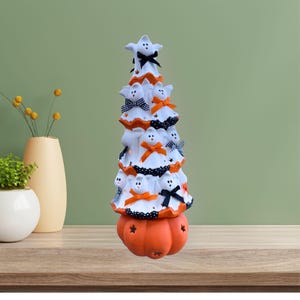 May include: A Halloween-themed decorative tree with a pumpkin base. The white tree is adorned with ghost figures, orange and black bows, and a star on top. The base is a carved orange pumpkin with star cutouts.