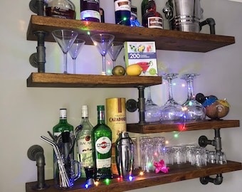 May include: A wooden shelf unit with black metal pipes, holding various bottles of liquor, glasses, and bar accessories. The shelf is decorated with fairy lights and a cocktail recipe book.