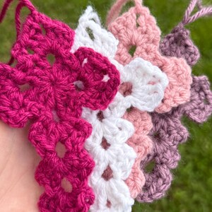 May include: Close-up of handmade crochet flowers in shades of fuchsia, white, light pink, and mauve. The crochet work features a detailed floral design, with visible yarn texture and openwork patterns. The flowers are arranged in a layered, overlapping fashion.