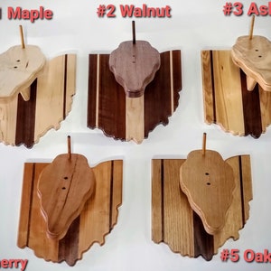 May include: Five wooden Ohio state-shaped plaques with different wood types. Each plaque has a small wooden cutout of the state of Ohio mounted on top. The wood types are Maple, Walnut, Ash, Cherry, and Oak. The plaques are numbered 1 through 5.