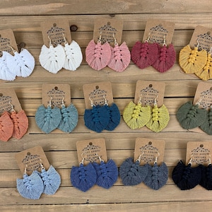 May include: A set of 15 pairs of handmade tassel earrings in various colors, including white, pink, red, yellow, orange, green, blue, and black. Each pair is made with a gold-colored hook and a heart-shaped tassel. The earrings are displayed on a wooden surface with small cards that say "Foxy Arrow Designs".