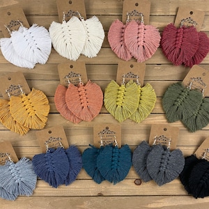 May include: Assortment of handmade macrame earrings in various colors, including white, pink, red, yellow, orange, green, blue, gray, and black. The earrings are all in the shape of a leaf and have a fringe detail.
