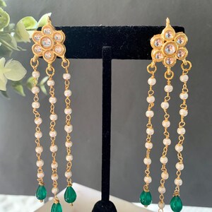 May include: Gold earrings with a floral design and white pearl chains. The earrings have green gemstone drops at the end of the chains.