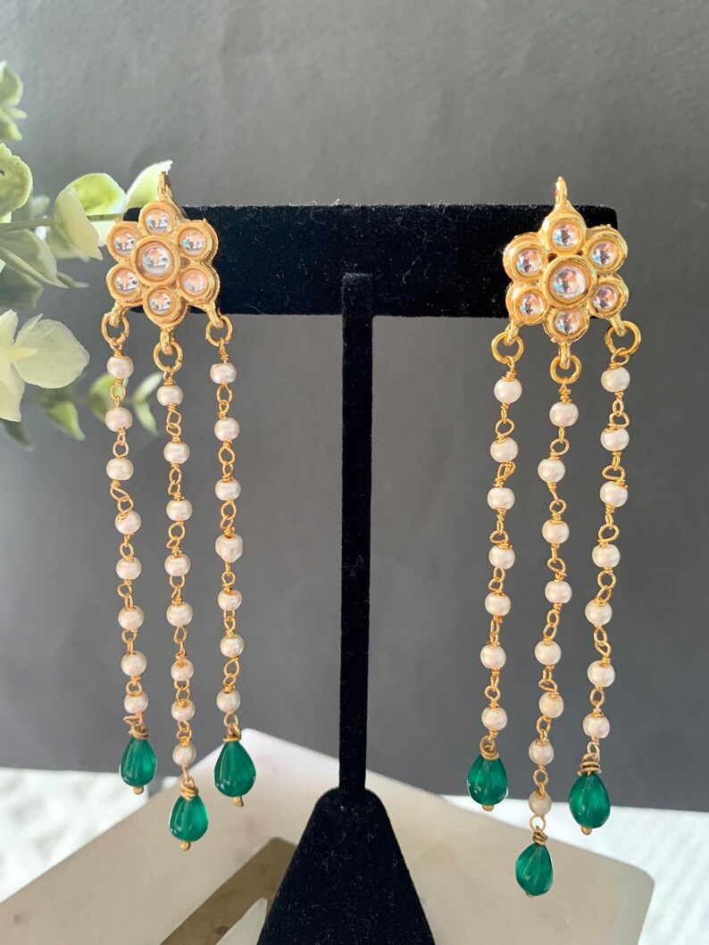 May include: Gold earrings with a floral design and white pearl chains. The earrings have green gemstone drops at the end of the chains.