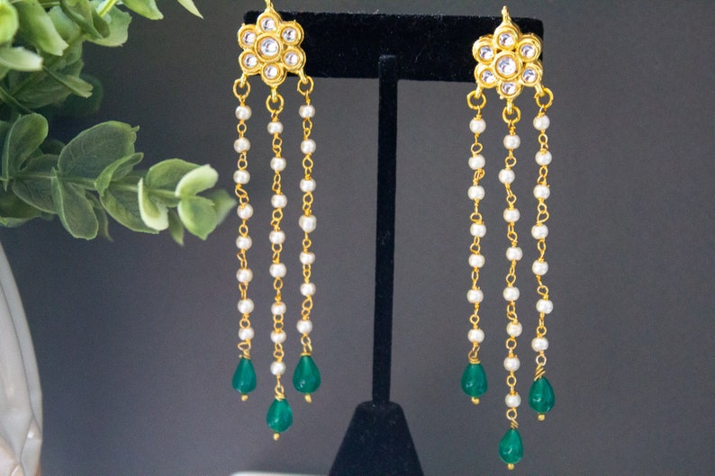 May include: Gold earrings with a floral design and white pearl chains, ending with green teardrop-shaped stones.