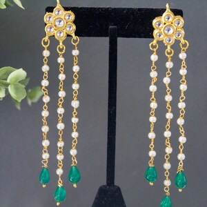 May include: Gold earrings with a floral design and white pearl beads hanging from the bottom. The earrings have three green teardrop-shaped stones at the end of the chains.
