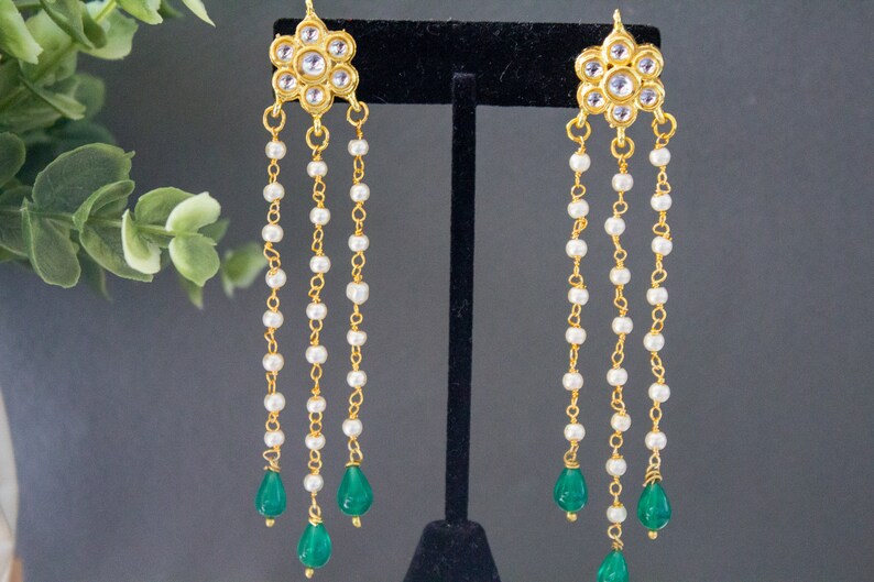 May include: Gold earrings with a floral design and white pearl beads hanging from the bottom. The earrings have three green teardrop-shaped stones at the end of the chains.