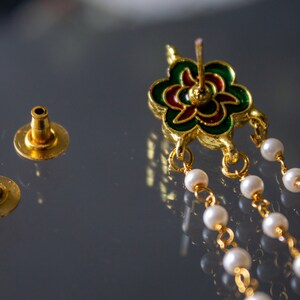 May include: A pair of gold earrings with a green, red and gold flower design. The earrings have a chain of white pearls hanging from the flower.