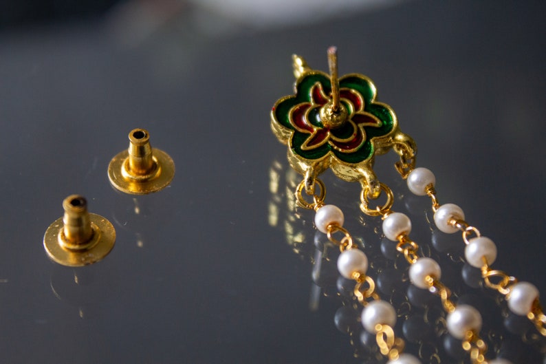 May include: A pair of gold earrings with a green, red and gold flower design. The earrings have a chain of white pearls hanging from the flower.