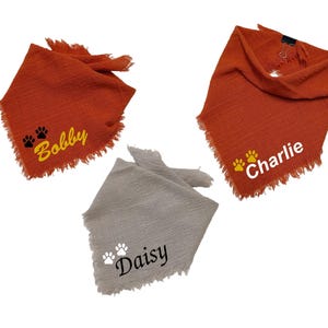 May include: Three personalized dog bandanas in shades of orange and gray. The orange bandanas have the names "Bobby" and "Charlie" embroidered with paw prints. The gray bandana is embroidered with the name "Daisy" and paw prints. All bandanas have a fringed edge.