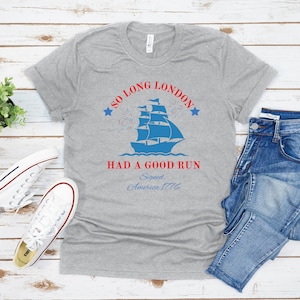 May include: A gray t-shirt with a blue and red graphic design. The graphic features a ship sailing away from a city skyline with fireworks in the background. The text on the shirt reads "So Long London Had A Good Run Signed, America 1776".