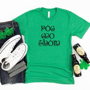 May include: Green t-shirt with the Irish phrase "Póg mo thóin" printed in black. The shirt is laid flat on a white background with a pair of jeans, white sneakers, and two bottles of Guinness.