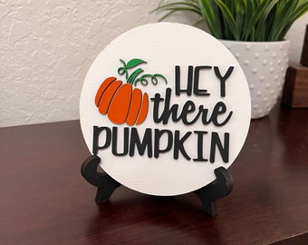May include: A white round wooden sign with a black stand. The sign has a pumpkin with a green stem and the words "Hey there pumpkin" in black lettering.