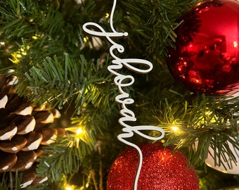 May include: White wooden Christmas ornament with the word "Jehovah" written in cursive script. The ornament is hanging on a green Christmas tree with red and white ornaments.