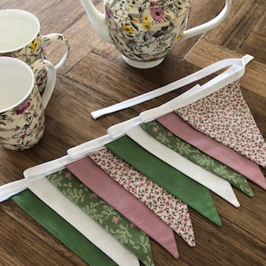 May include: A white fabric bunting with green and pink floral patterned flags. The bunting is strung on a white ribbon and is hanging on a wooden surface.