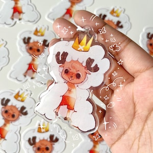 Op de afbeelding: Een wit, pluche personage met een kroon en een rode broek, dat een wit, pluche jasje met glitter draagt, is afgebeeld op een transparante acryl hanger. De hanger wordt in een hand gehouden.