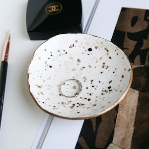 Op de afbeelding: Een witte keramische ringenschotel met gouden rand en een gespikkelde afwerking. Een zilveren ring ligt in de schotel.
