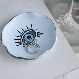 Può includere: Un porta anelli in ceramica bianca con un design a occhio blu. L'occhio ha un contorno nero e ciglia. Due anelli d'argento sono posizionati nel piatto.