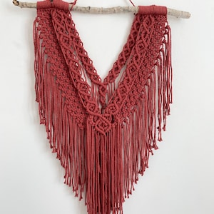 May include: A terracotta-colored macrame wall hanging with a V-shaped design and fringe. The hanging is attached to a natural branch.