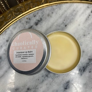 Puede incluir: Una lata de metal plateada con una etiqueta rosa que dice "Chaotically Curated Intensive Lip Balm". La etiqueta enumera los ingredientes: cera de abejas, aceite de babassu, lanolina, glicerina vegetal y aceite mineral. La lata contiene un bálsamo labial amarillo y está etiquetada con 1.5 fl oz.