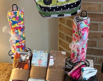 May include: A set of colourful reusable kitchen towels with various patterns, including a snowman, a mushroom, and a watermelon. The towels are folded and displayed on a table with a basket of additional towels.