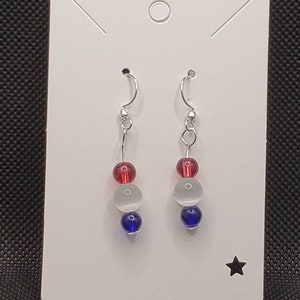 May include: A pair of silver dangle earrings with red, white, and blue glass beads. The earrings are displayed on a white card with a star.