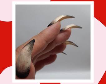 May include: Close-up of a hand with long, sharp, fake nails. The nails are painted with a black and beige ombre effect.