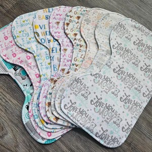 May include: A collection of contoured burp cloths featuring various printed patterns including text such as I love my mommy, I love my daddy, and Love you to the moon and back, alongside illustrations of teddy bears, elephants, and cows in pastel colors.