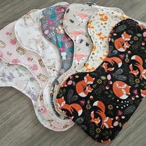 May include: A set of seven fabric burp cloths with various woodland animal prints, including foxes, deer, and owls. The cloths are in shades of pink, white, beige, gray, and black.