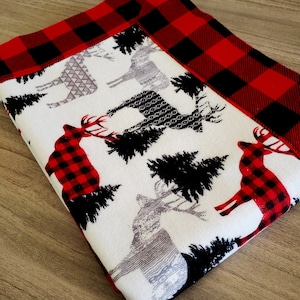 May include: A white blanket with a red and black buffalo plaid border. The blanket features a repeating pattern of gray and black deer and black pine trees.