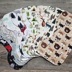 May include: A collection of baby burp cloths with various woodland animal and nature-themed patterns. Designs include foxes, bears, deer, trees, and camping motifs. The cloths are in shades of gray, black, red, and beige, arranged in a fan shape.