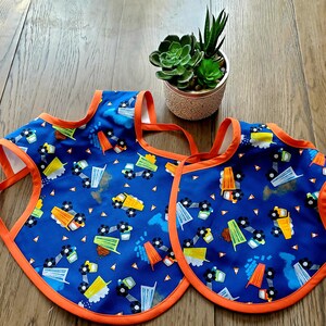 May include: Two blue bibs with an orange trim and a pattern of construction trucks on a wooden surface.