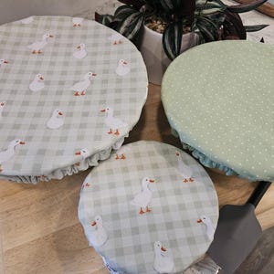 May include: Three round fabric bowl covers with elastic edges. Two covers feature a light green and white gingham pattern with a repeating white goose print. One cover features a solid light green fabric with a small white polka dot pattern.