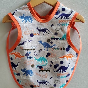 May include: A gray and orange baby bib with a dinosaur print. The bib features various dinosaurs, including Triceratops, Brachiosaurus, and Stegosaurus. The bib has a rounded bottom and an orange trim.