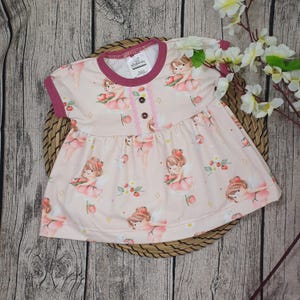 Könnte beinhalten: Hellrosa Babykleid mit einem Muster aus Cartoon-Feen und Erdbeeren. Das Kleid hat kurze Ärmel und einen runden Halsausschnitt mit dunkelrosa Besatz. Es hat eine Knopfleiste vorne und einen gerafften Rock.