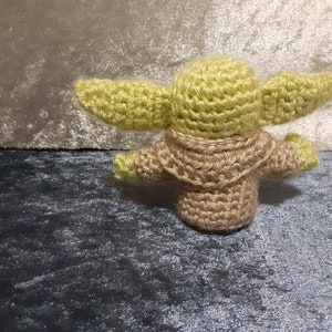 May include: A crocheted green and brown Yoda doll. The doll is standing on a blue and grey surface.