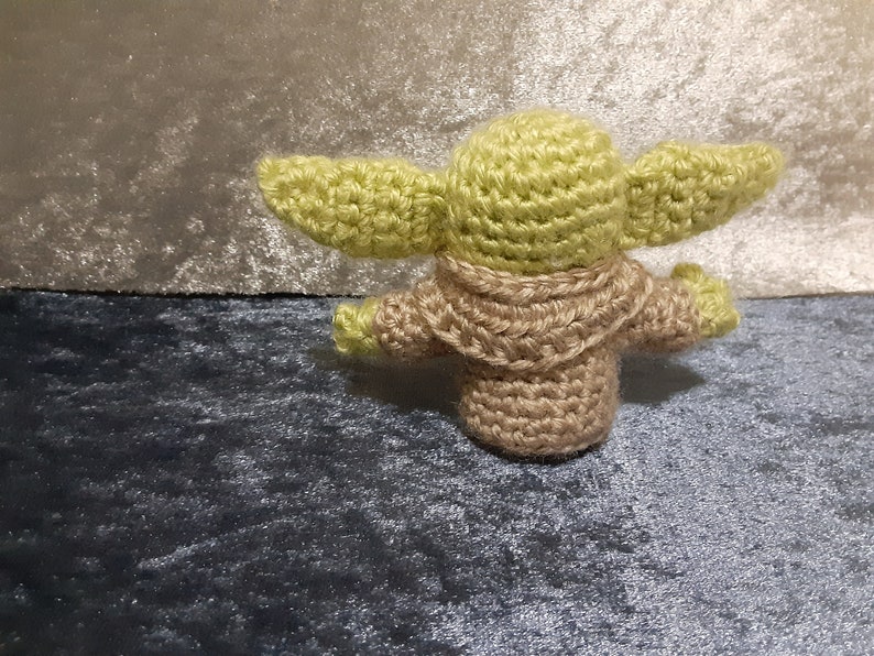 May include: A crocheted green and brown Yoda doll. The doll is standing on a blue and grey surface.