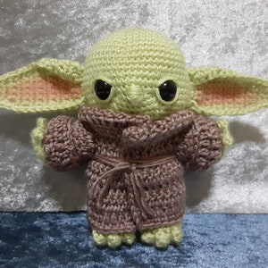 May include: A crocheted plush toy of a green character with large ears, wearing a purple robe. The character has black eyes and a serious expression.