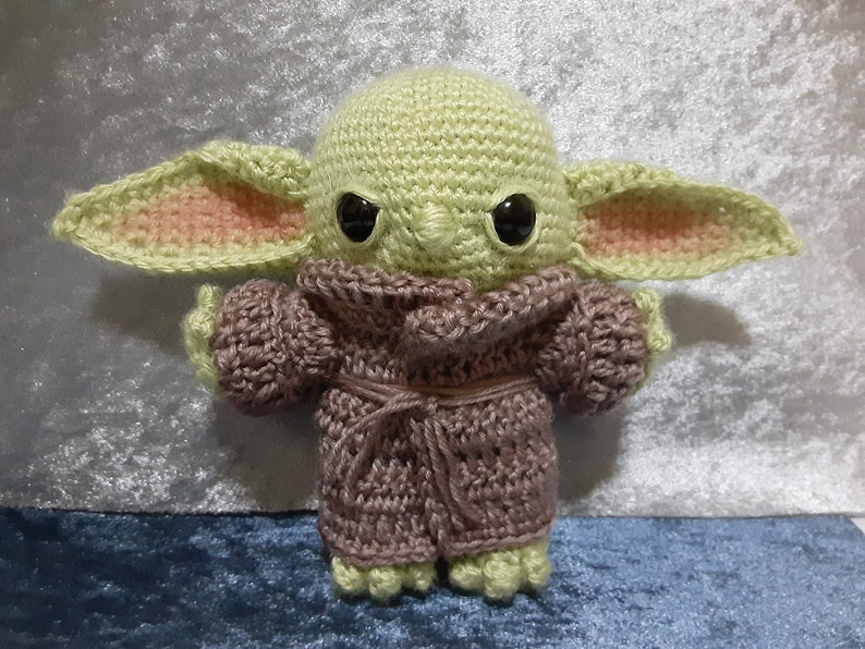 May include: A crocheted plush toy of a green character with large ears, wearing a purple robe. The character has black eyes and a serious expression.