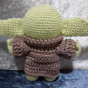 May include: A crocheted plush toy of a green alien character wearing a brown robe. The toy has large ears and a round head.