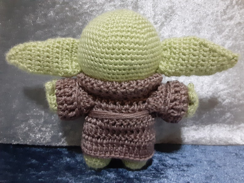 May include: A crocheted plush toy of a green alien character wearing a brown robe. The toy has large ears and a round head.
