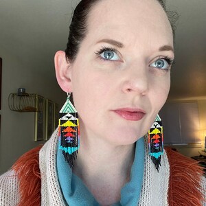 May include: A woman wearing a blue turtleneck sweater and a white and orange cardigan is wearing a pair of long, colorful beaded earrings. The earrings are black, white, green, blue, orange, yellow, and red.